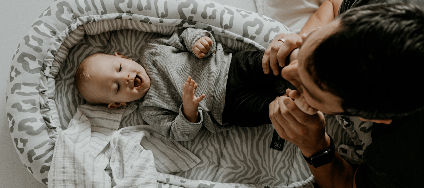 Baby liegt in Babynestchen Leo Grau und spielt mit Papa